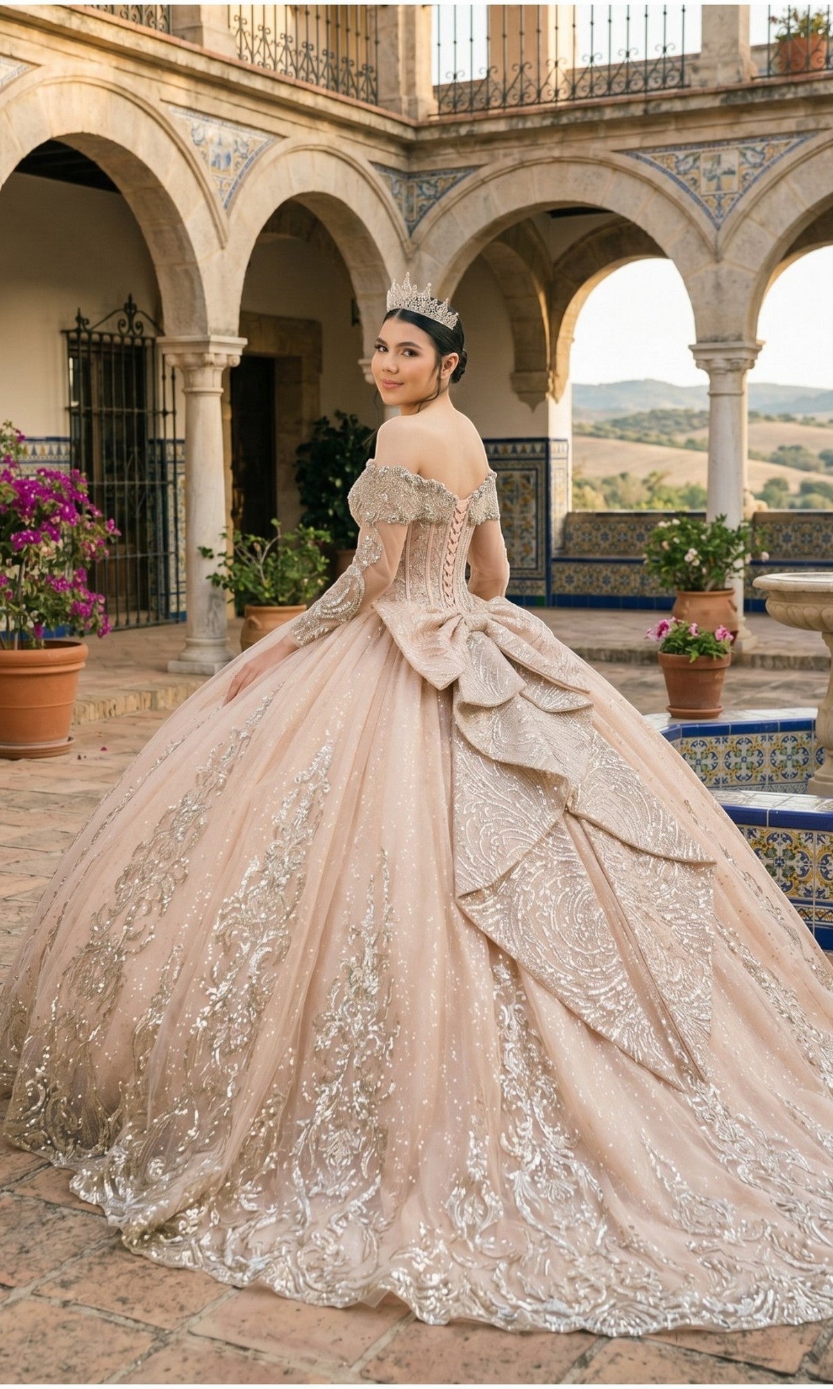 Wearing the Quinceanera Dress 8600JQ by Cinderella Couture, a young woman stands in a courtyard with arched columns, smiling over her shoulder in an ornate gold ball gown featuring a sparkling finish, off-the-shoulder neckline, and elegant corset bodice.